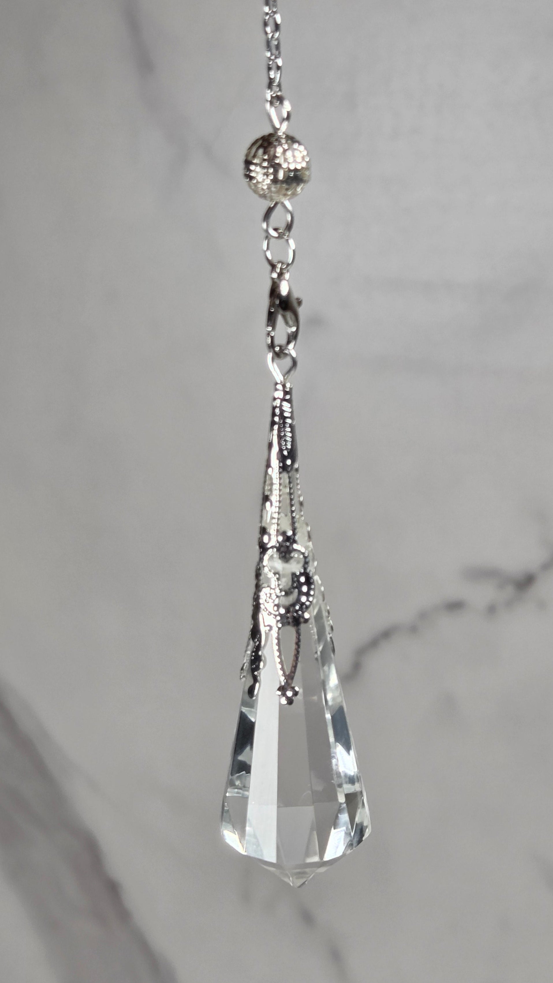 A silver-toned chain accessory with a faceted clear crystal pendant, displayed horizontally against a light gray marble background.