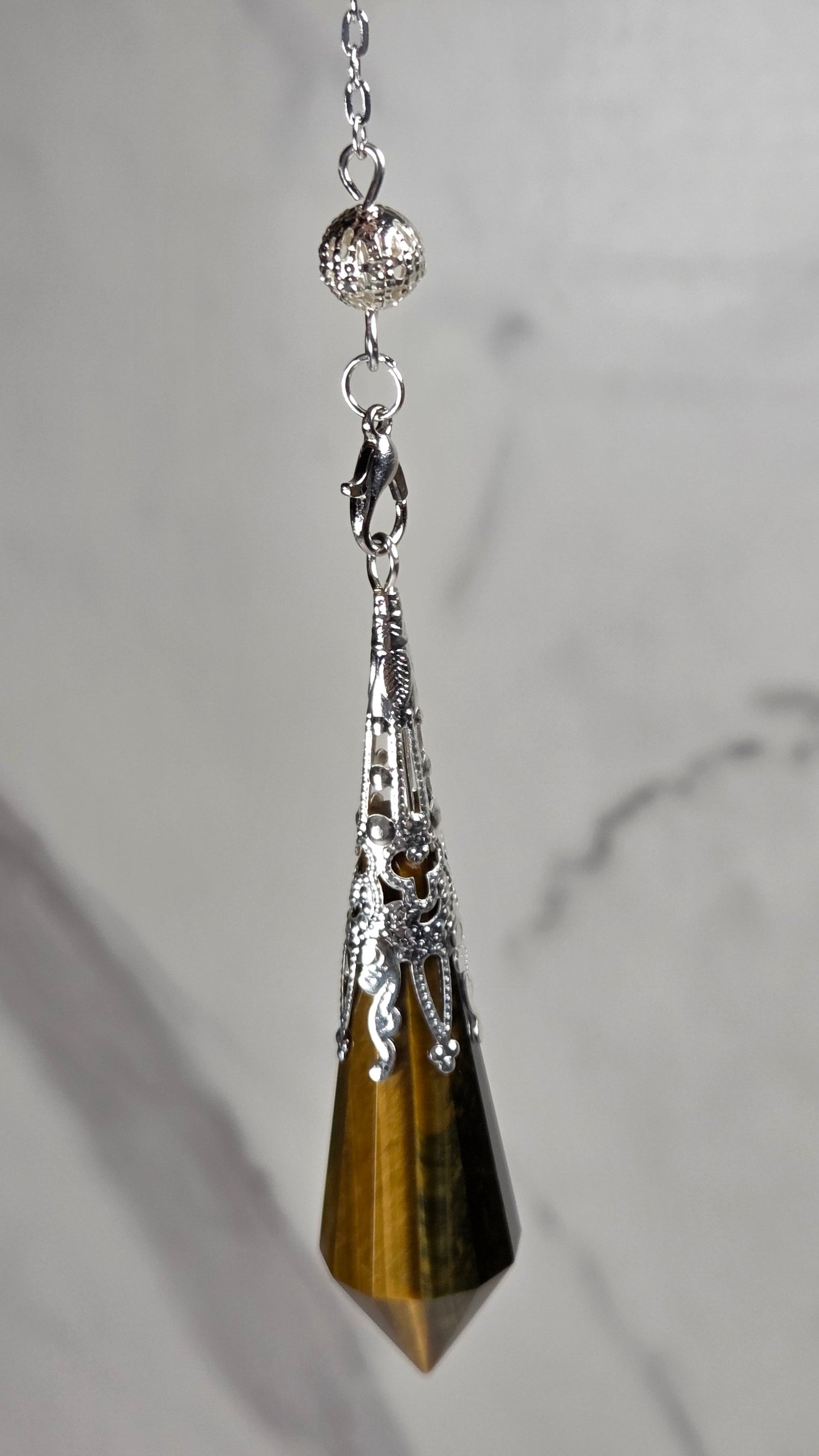 A silver-toned pendant pendulum featuring a faceted tiger's eye crystal point, resting on a light gray marble surface.