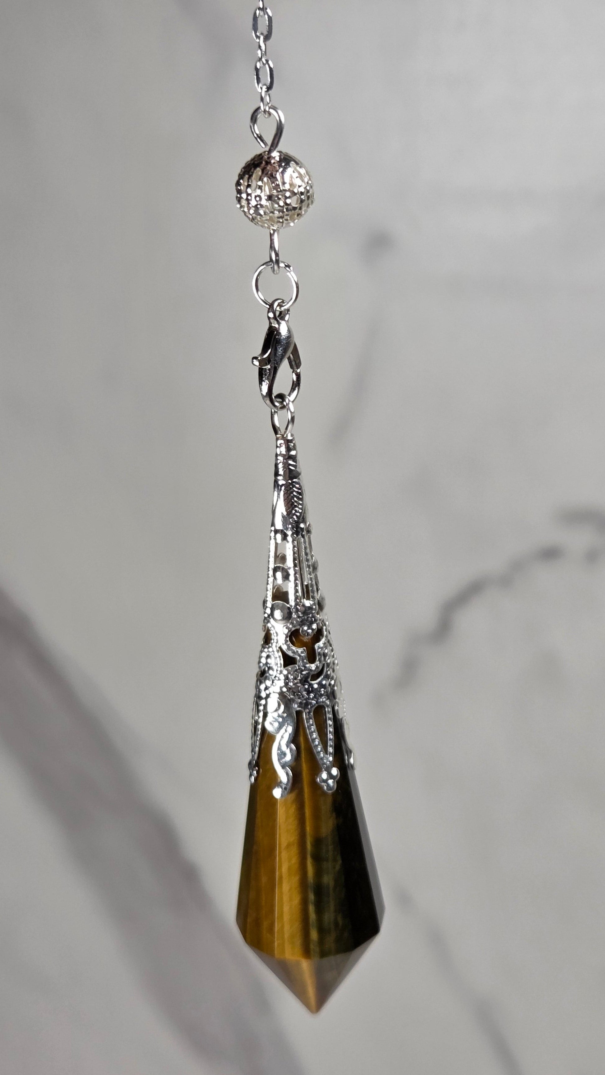 A silver-toned pendant pendulum featuring a faceted tiger's eye crystal point, resting on a light gray marble surface.