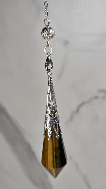 A silver-toned pendant pendulum featuring a faceted tiger's eye crystal point, resting on a light gray marble surface.