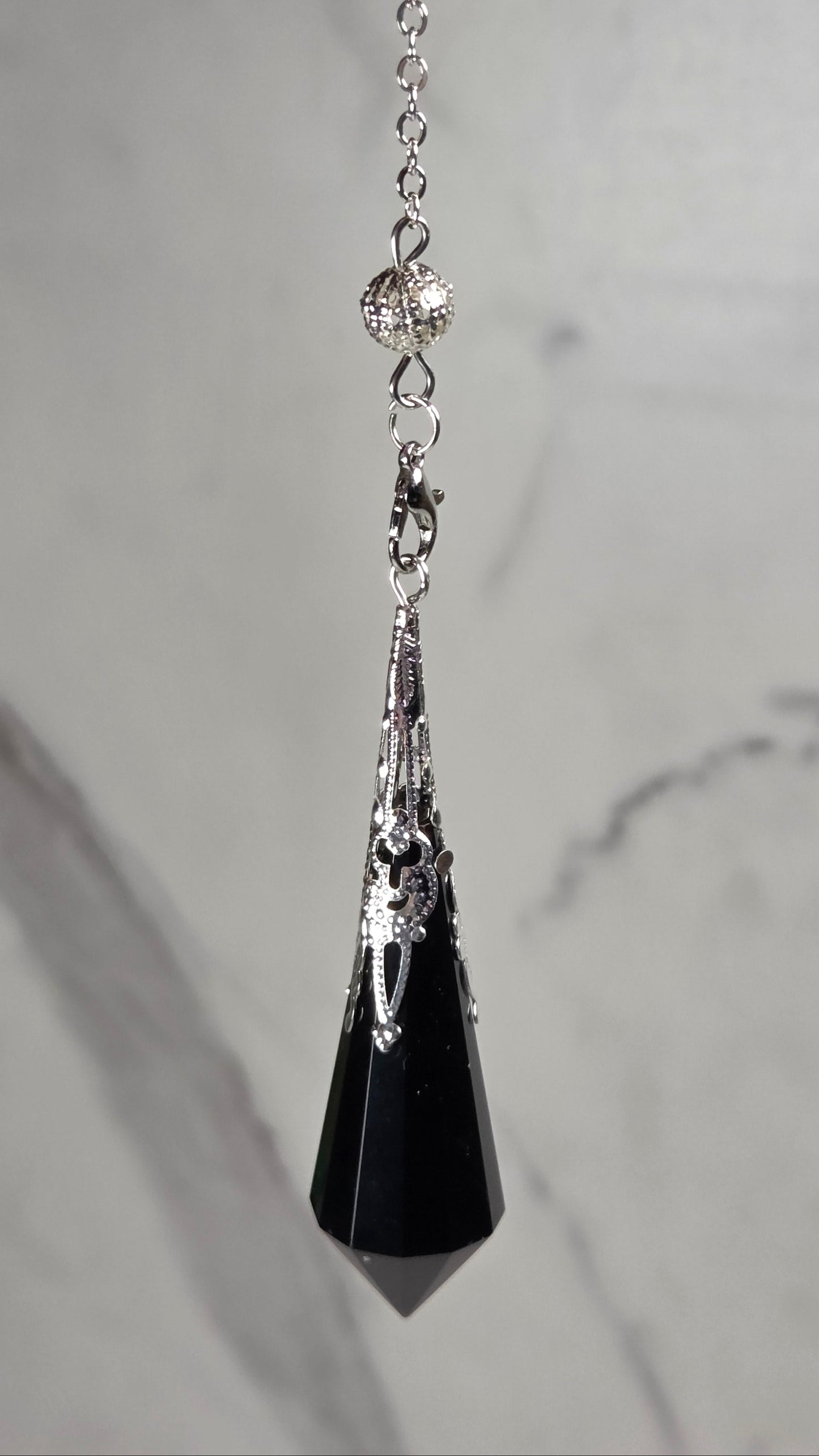 A silver-toned chain pendulum with a black faceted crystal pendant, displayed on a light gray marble surface.