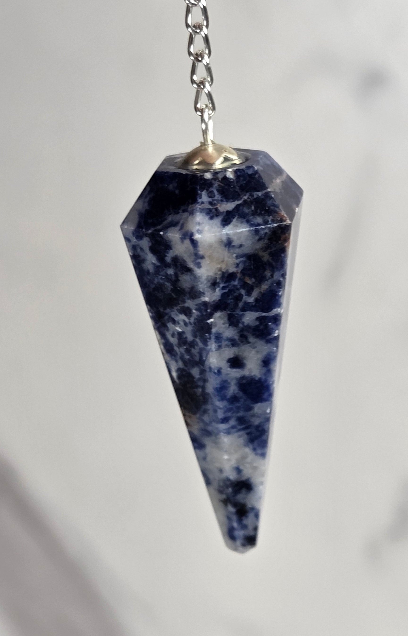A hand holding a pointed blue and white speckled stone pendant on a silver chain against a light marble background.