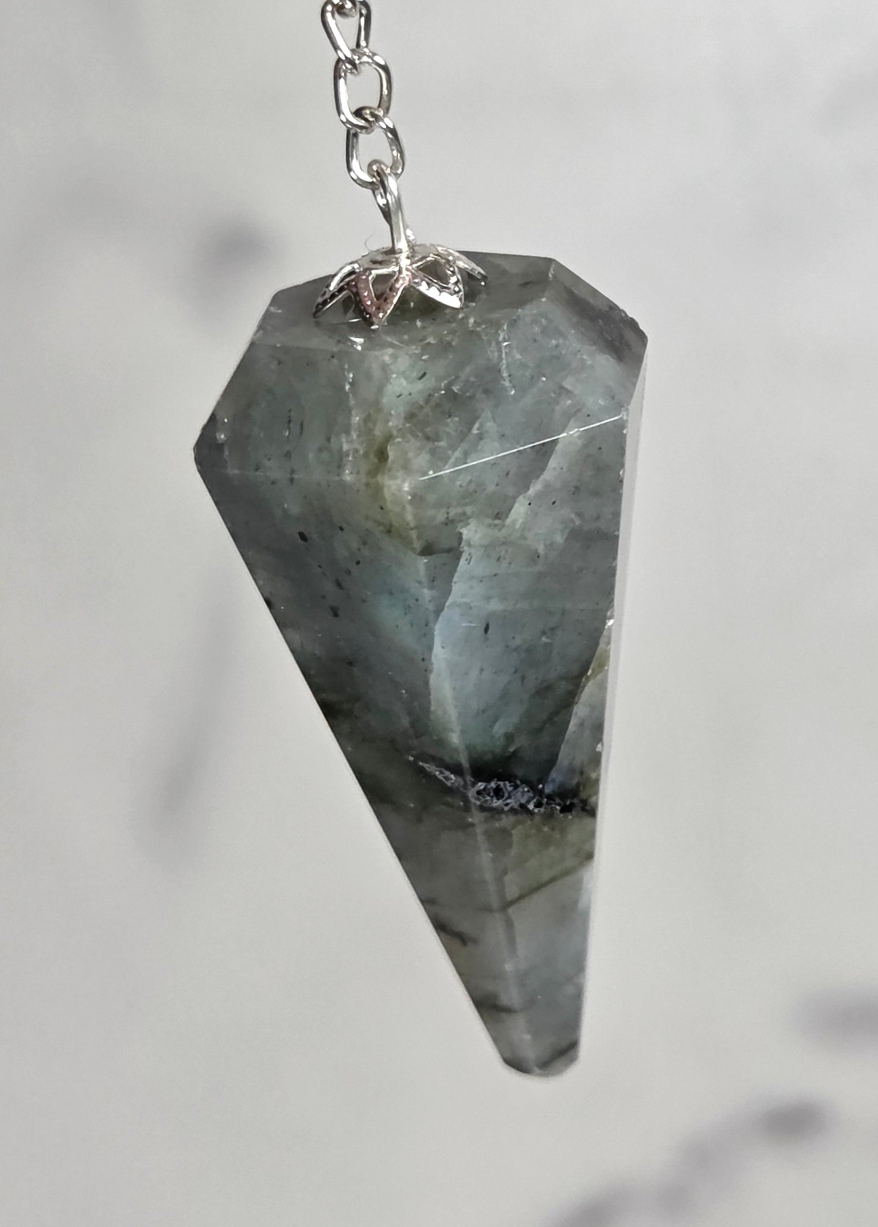A hand holding a gray labradorite cone pendant on a silver chain against a light gray marble background.