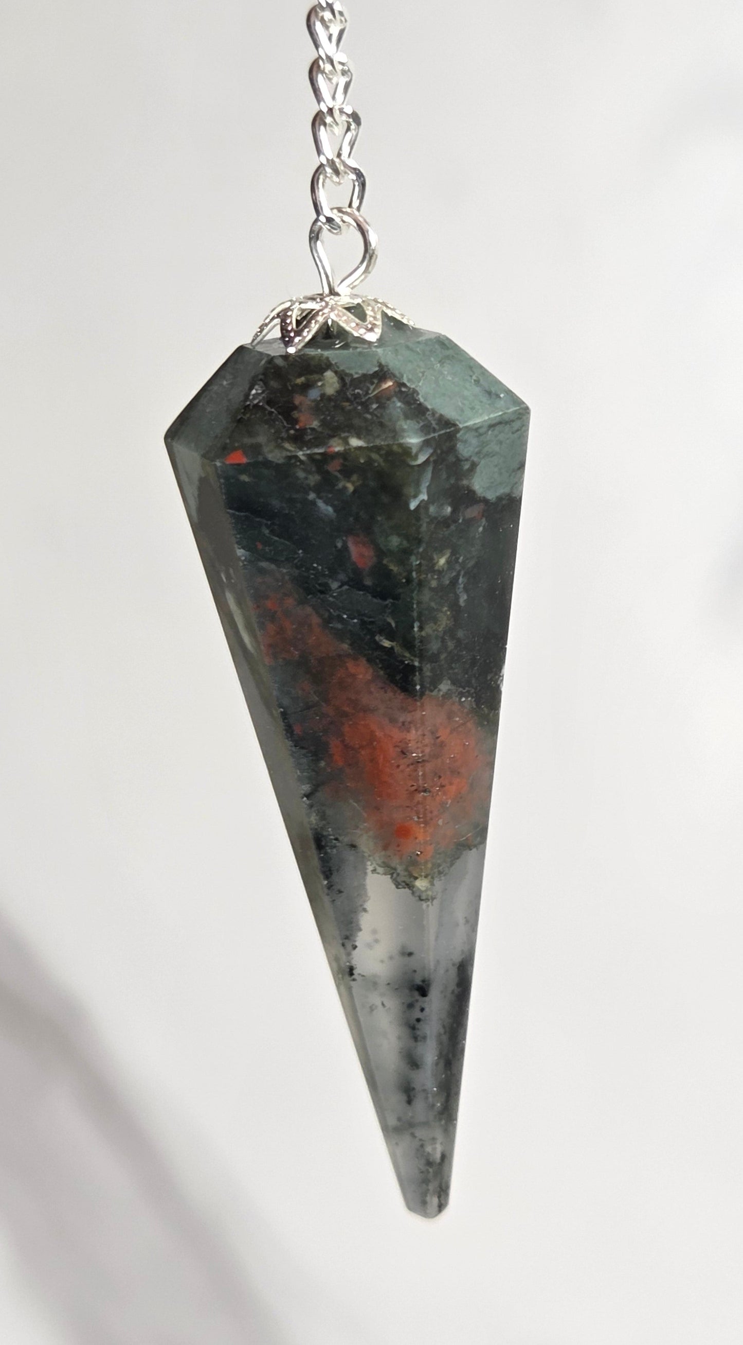 A faceted pointed crystal pendant pendulum in dark gray with orange and green inclusions, attached to a silver chain against a light gray marble background.