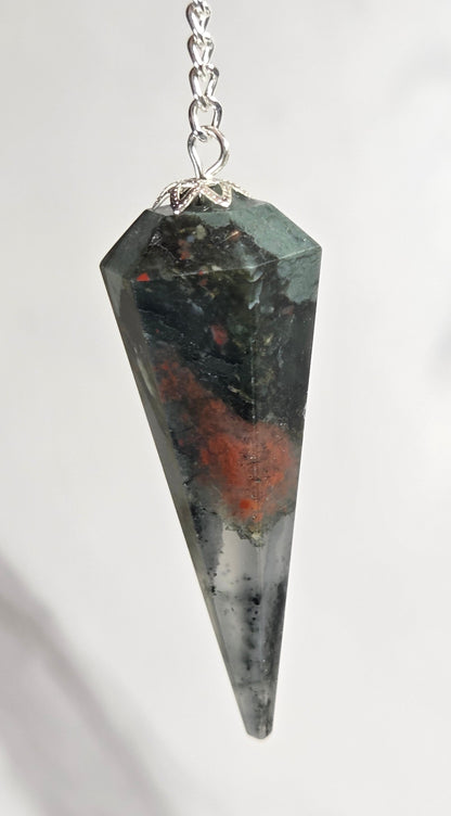 A faceted pointed crystal pendant pendulum in dark gray with orange and green inclusions, attached to a silver chain against a light gray marble background.