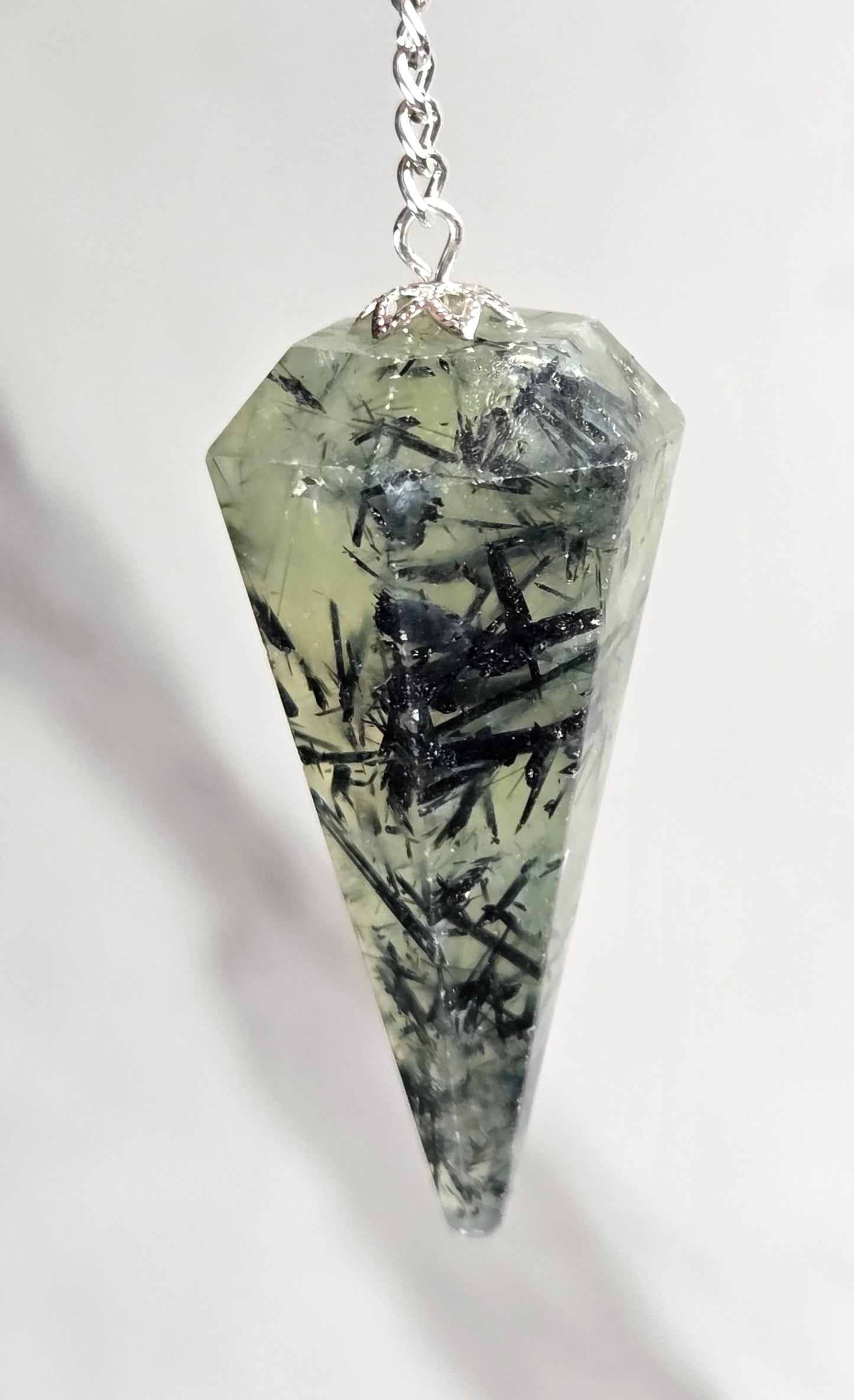 A pointed crystal pendant pendulum with a silver chain, held by a person's fingers against a light marble background. The crystal is pale greenish-gray with prominent black mineral inclusions.