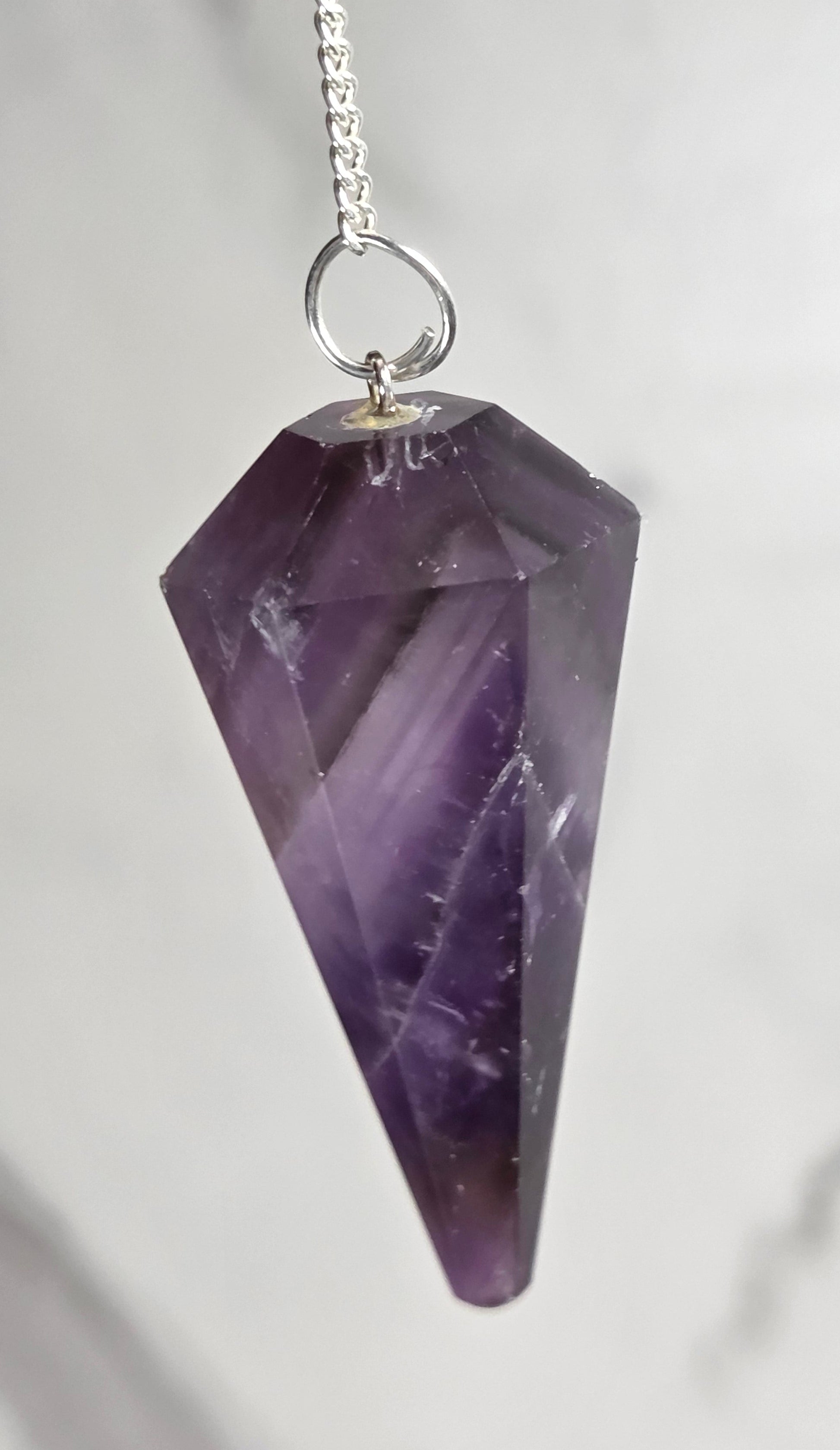 A faceted purple amethyst crystal pendant on a silver chain, photographed close-up against a light gray marble background.