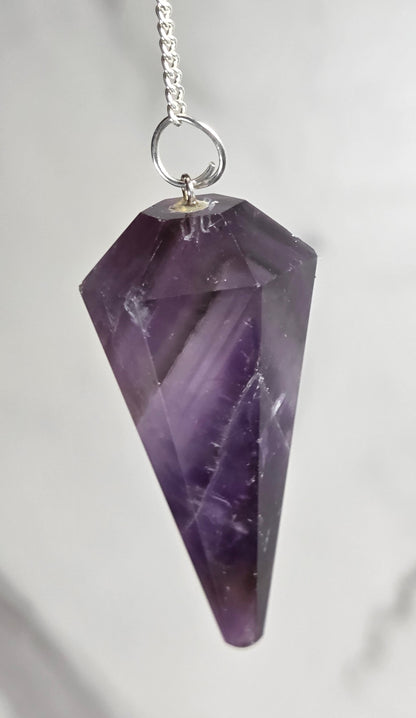 A faceted purple amethyst crystal pendant on a silver chain, photographed close-up against a light gray marble background.