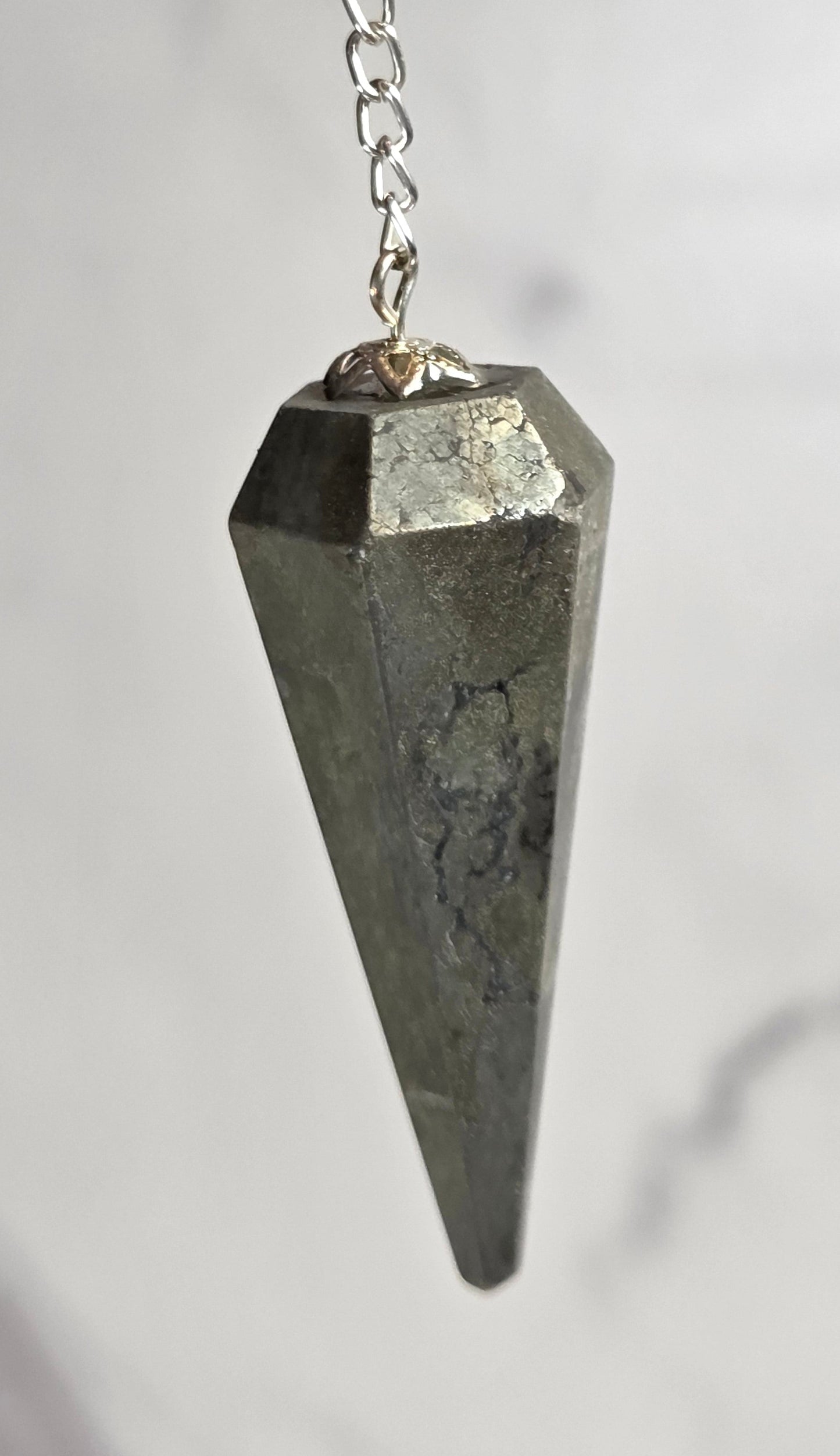 A metallic pyramid-shaped weight on a short silver chain, held by a finger against a light gray marble background.