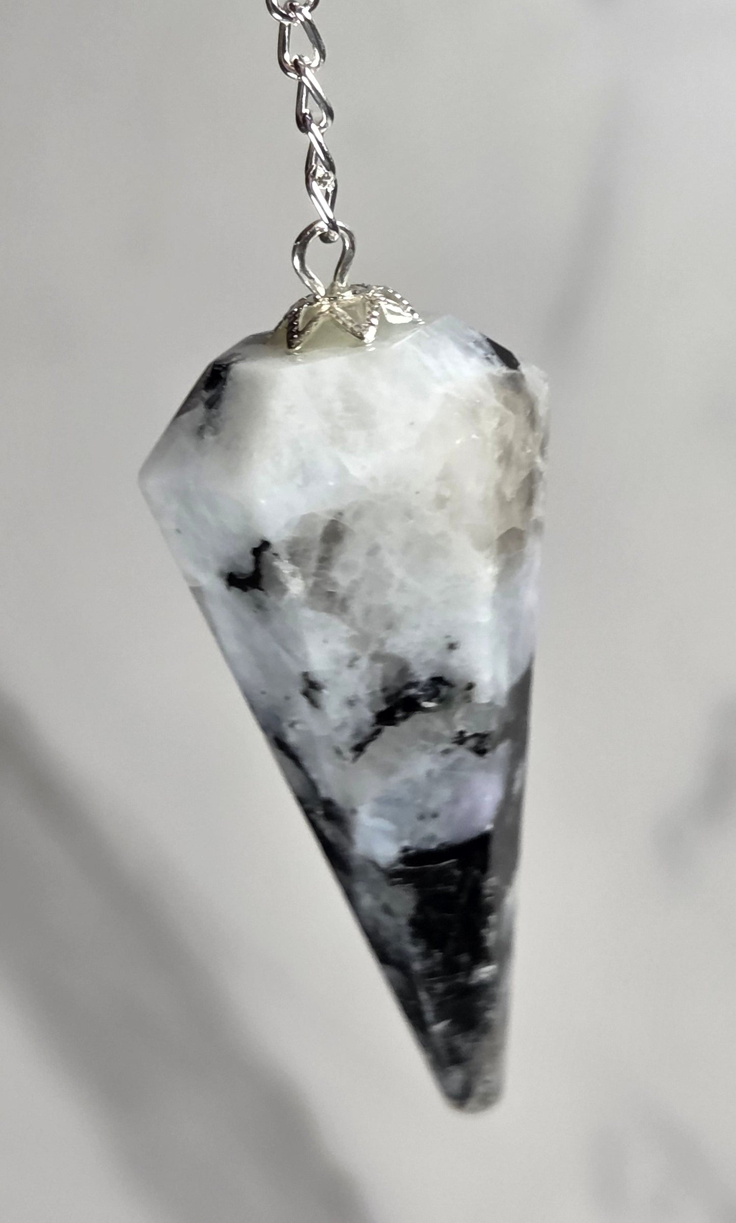 A silver-toned metal chain pendulum  with a faceted cone-shaped crystal pendant, featuring a white and black marbled pattern, shown against a light gray marble background.