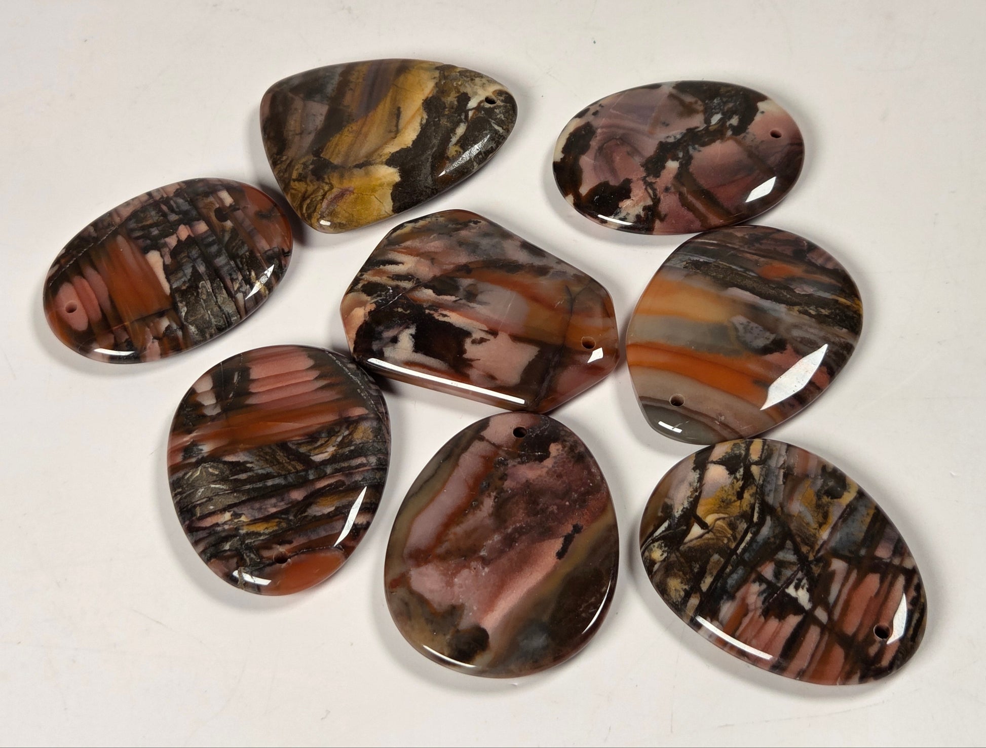 Seven polished cabochon gemstone pendants with natural agate-like banding in earthy brown, black, tan, and cream, arranged on a plain off-white background.