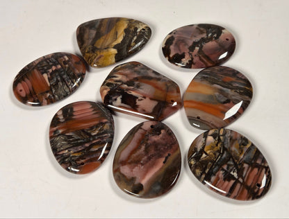 Seven polished cabochon gemstone pendants with natural agate-like banding in earthy brown, black, tan, and cream, arranged on a plain off-white background.