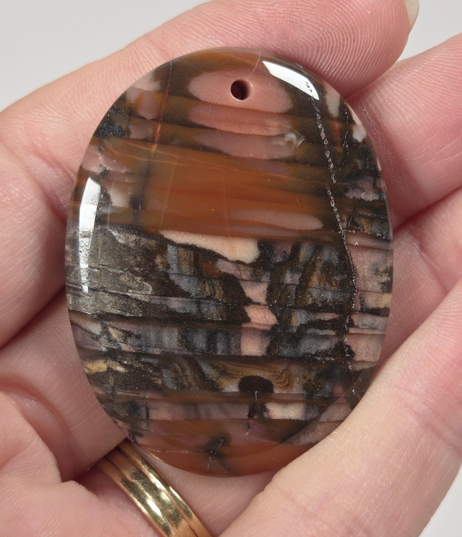 A hand holding an oval polished stone pendant with a natural wood grain pattern in shades of brown, black, and beige.