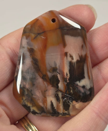 A polished stone pendant with a natural marbled pattern of brown, black, and cream, held in a person's hand.