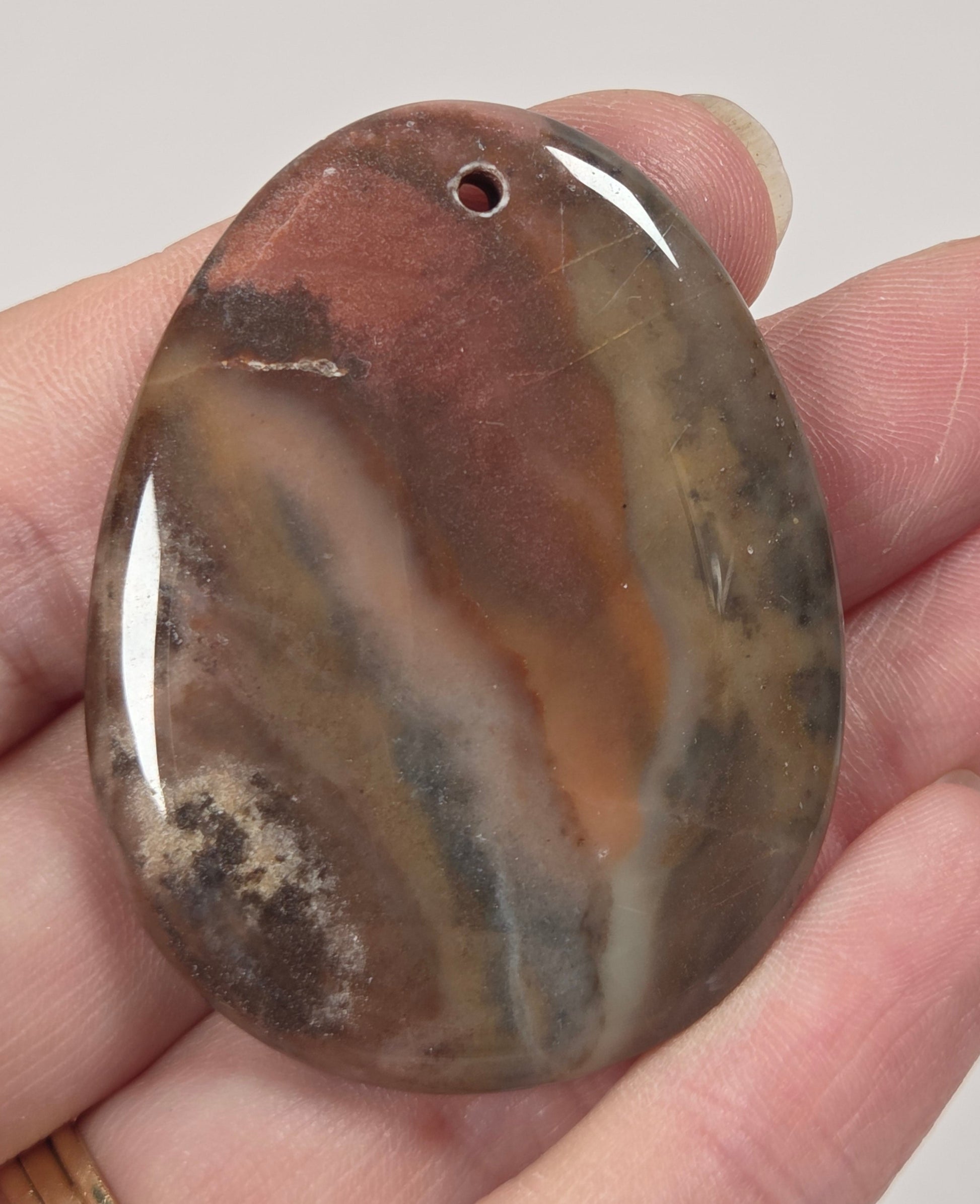 A hand holding a polished oval stone pendant with a pre-drilled hole, featuring natural banding in earthy brown, beige, gray, and black tones.