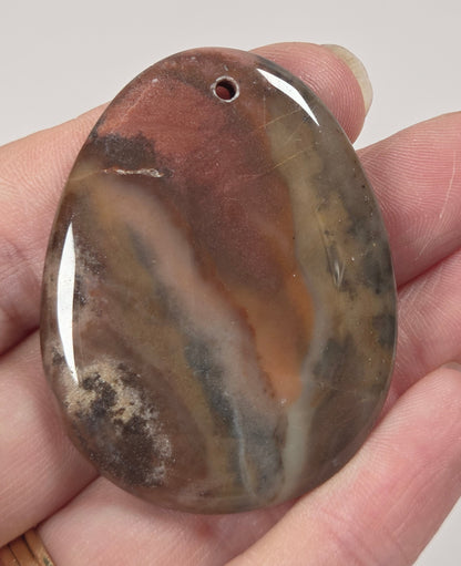 A hand holding a polished oval stone pendant with a pre-drilled hole, featuring natural banding in earthy brown, beige, gray, and black tones.