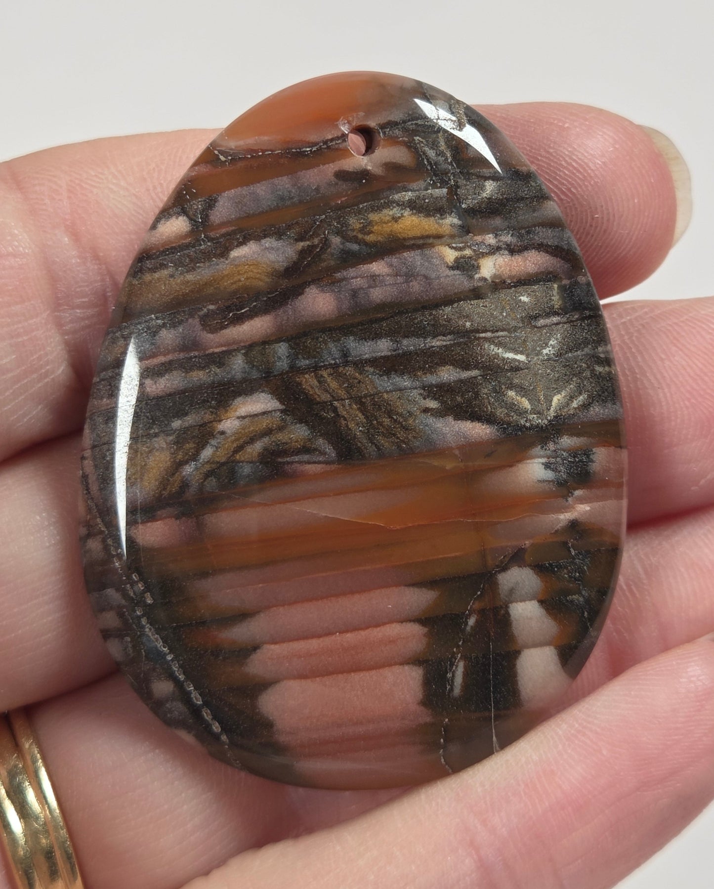 A hand holding a polished oval stone cabochon with a marbled pattern of brown, beige, and orange, with a small hole on one side.