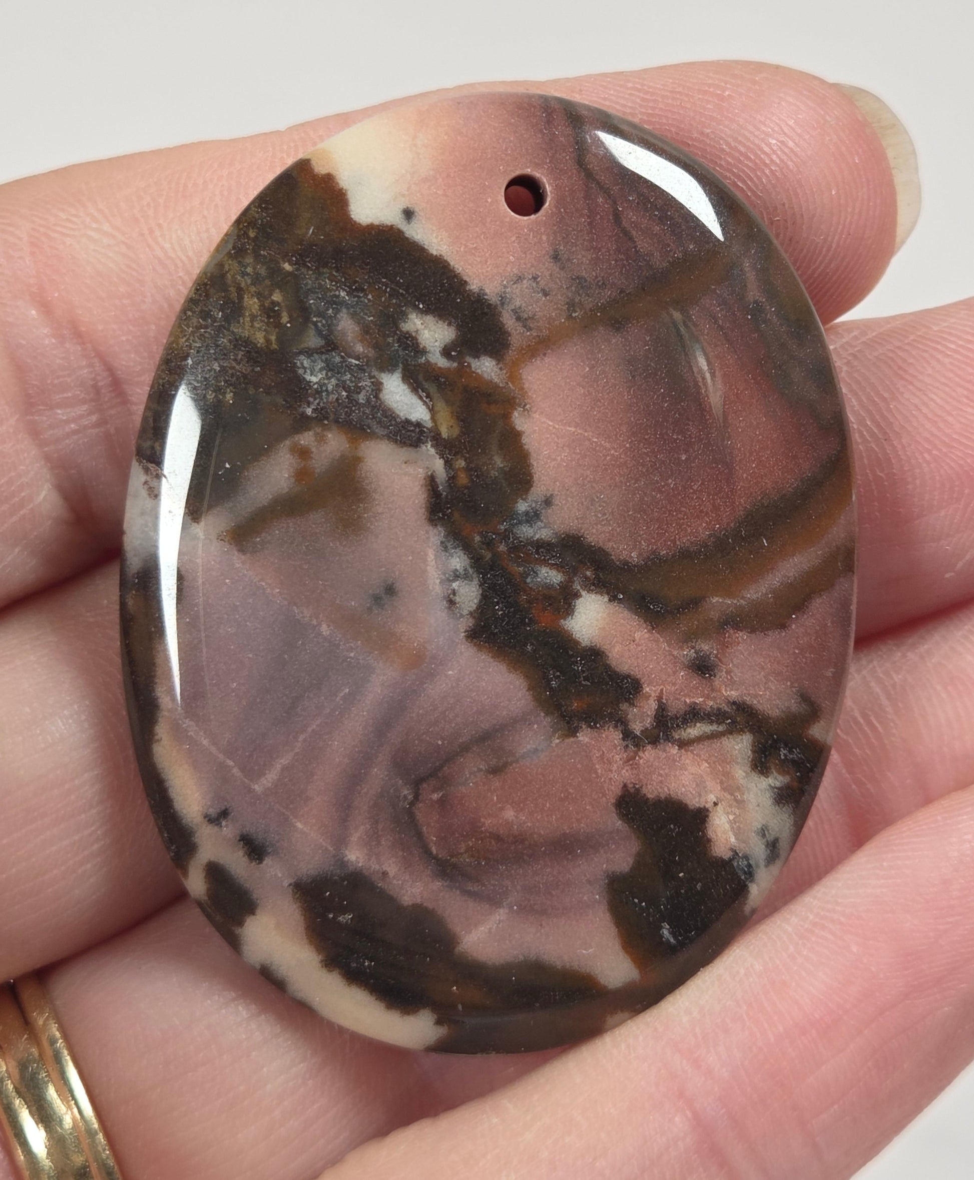 A polished oval agate stone cabochon with a pre-drilled hole, held in a person's hand. The stone features natural banding in shades of brown, beige, and black.