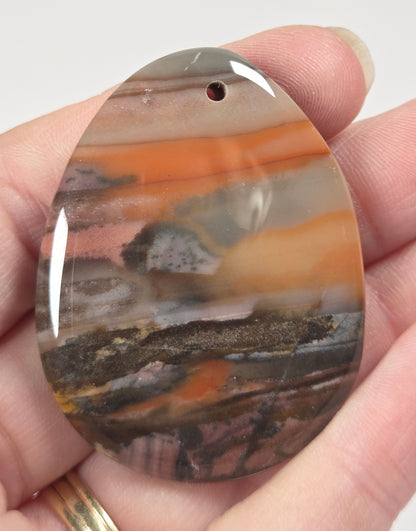 A hand holding a polished, oval-shaped agate stone cabochon pendant with a pre-drilled hole, showcasing its natural banding patterns in orange, brown, gray, and black.