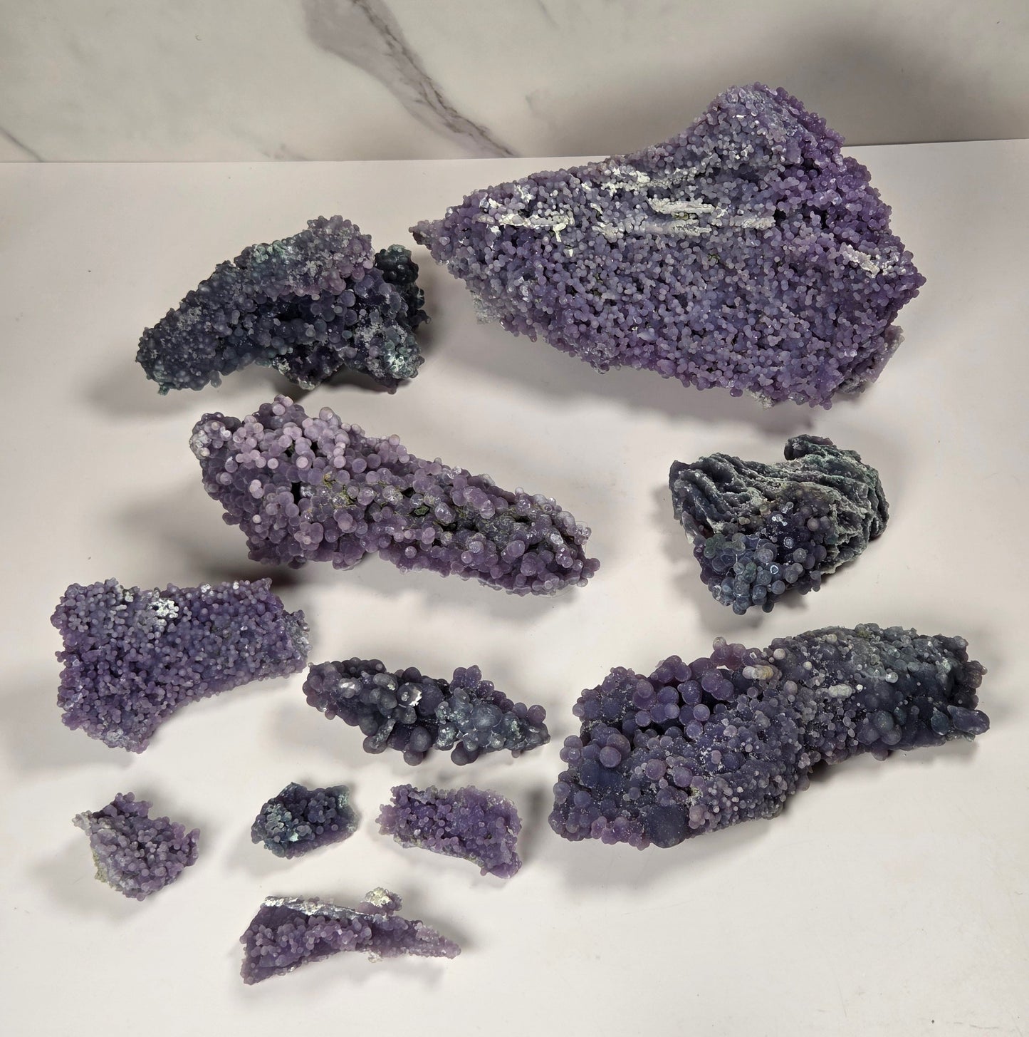 A collection of purple and blue-purple mineral specimens and crystal clusters arranged on a white surface with a white and gray marble-patterned background.