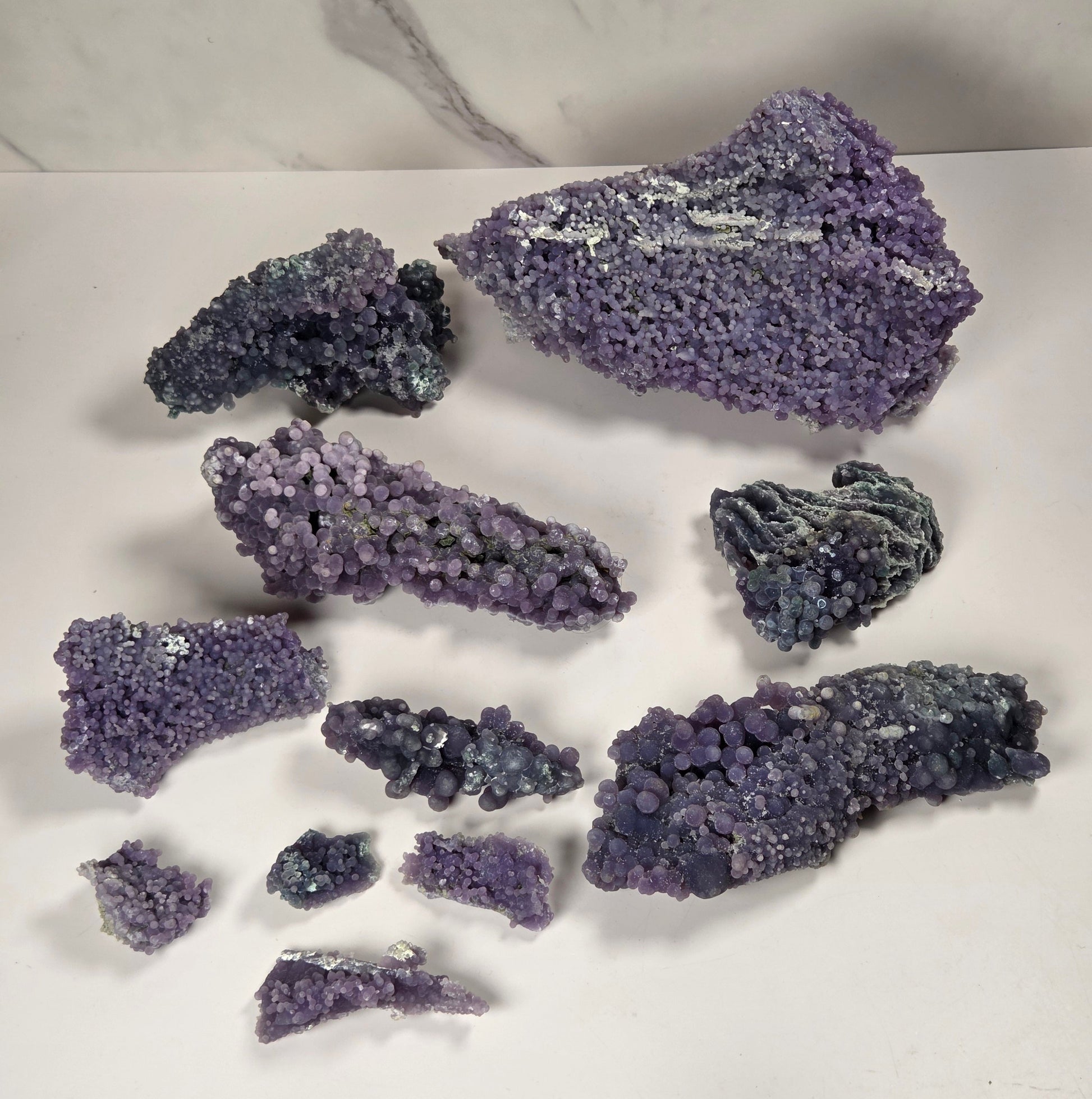 A collection of purple and blue-purple mineral specimens and crystal clusters arranged on a white surface with a white and gray marble-patterned background.