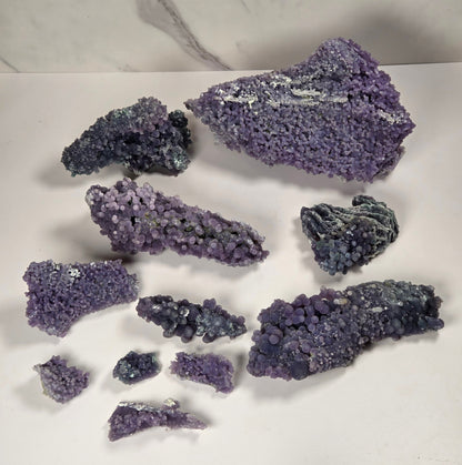 A collection of purple and blue-purple mineral specimens and crystal clusters arranged on a white surface with a white and gray marble-patterned background.