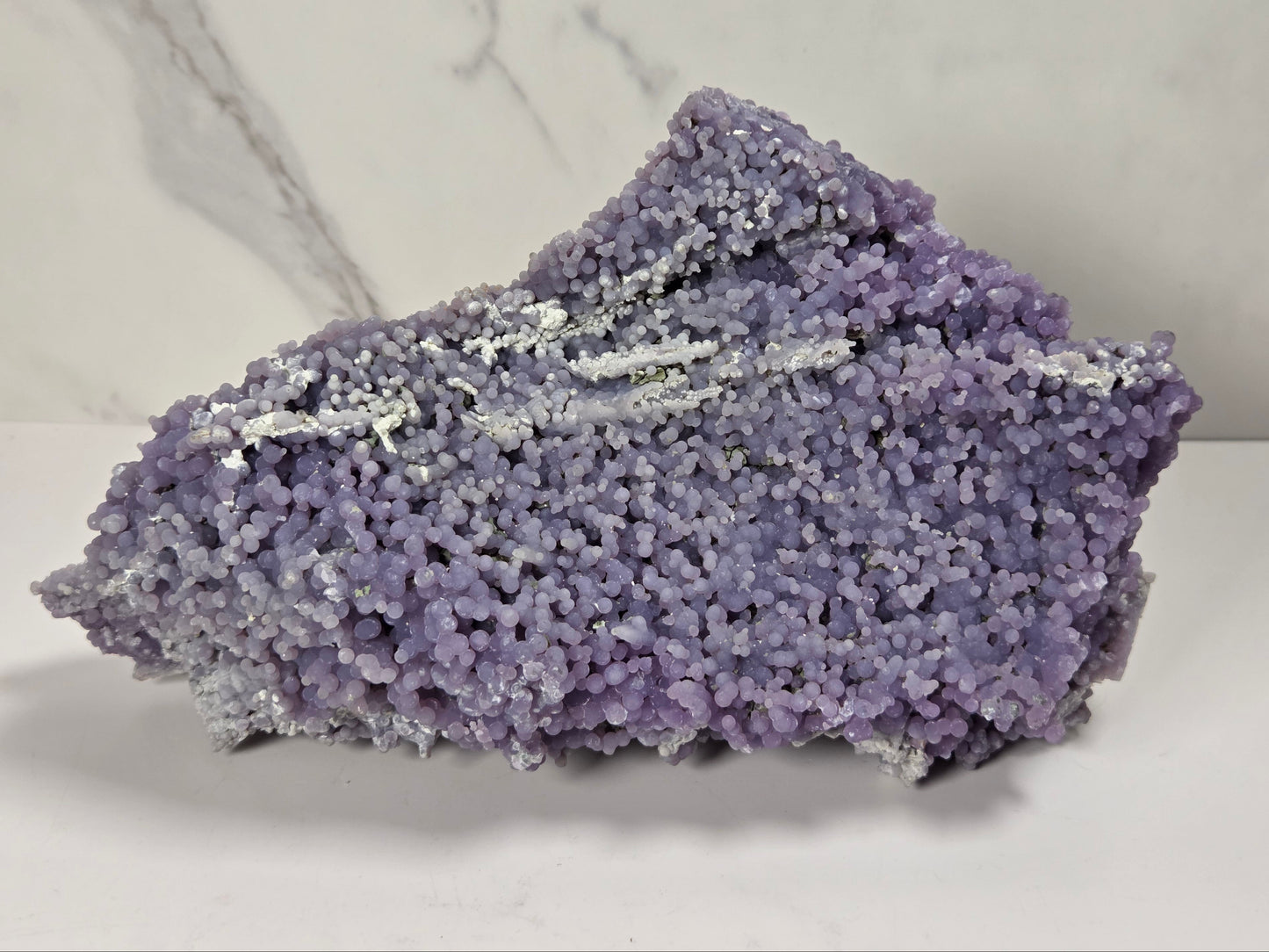 A purple and lavender crystal mineral specimen with dense, textured growth and small white crystalline inclusions, displayed on a white surface against a light gray marble-patterned background.