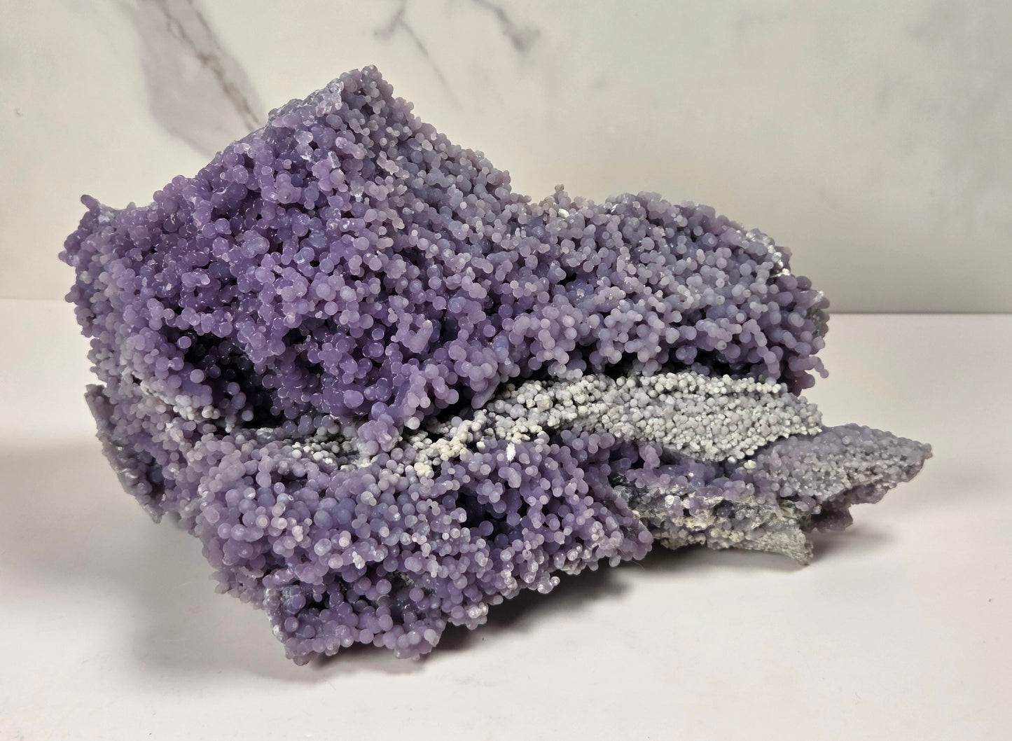 A purple and silver crystalline mineral specimen with a textured, coral-like surface, displayed on a white surface against a light gray marble-patterned background.