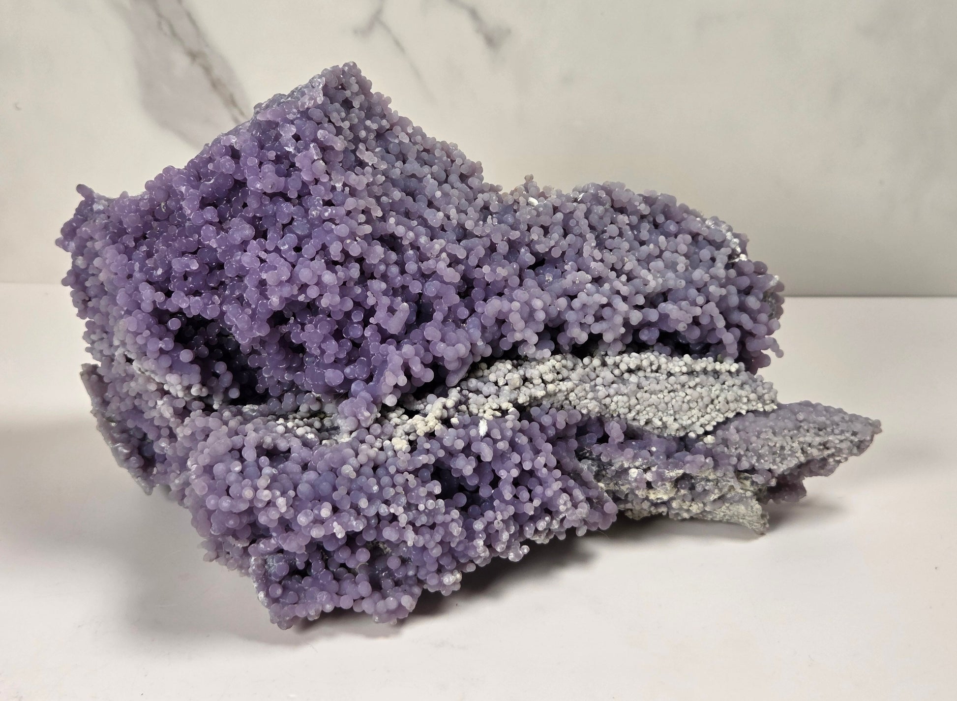 A purple and silver crystalline mineral specimen with a textured, coral-like surface, displayed on a white surface against a light gray marble-patterned background.
