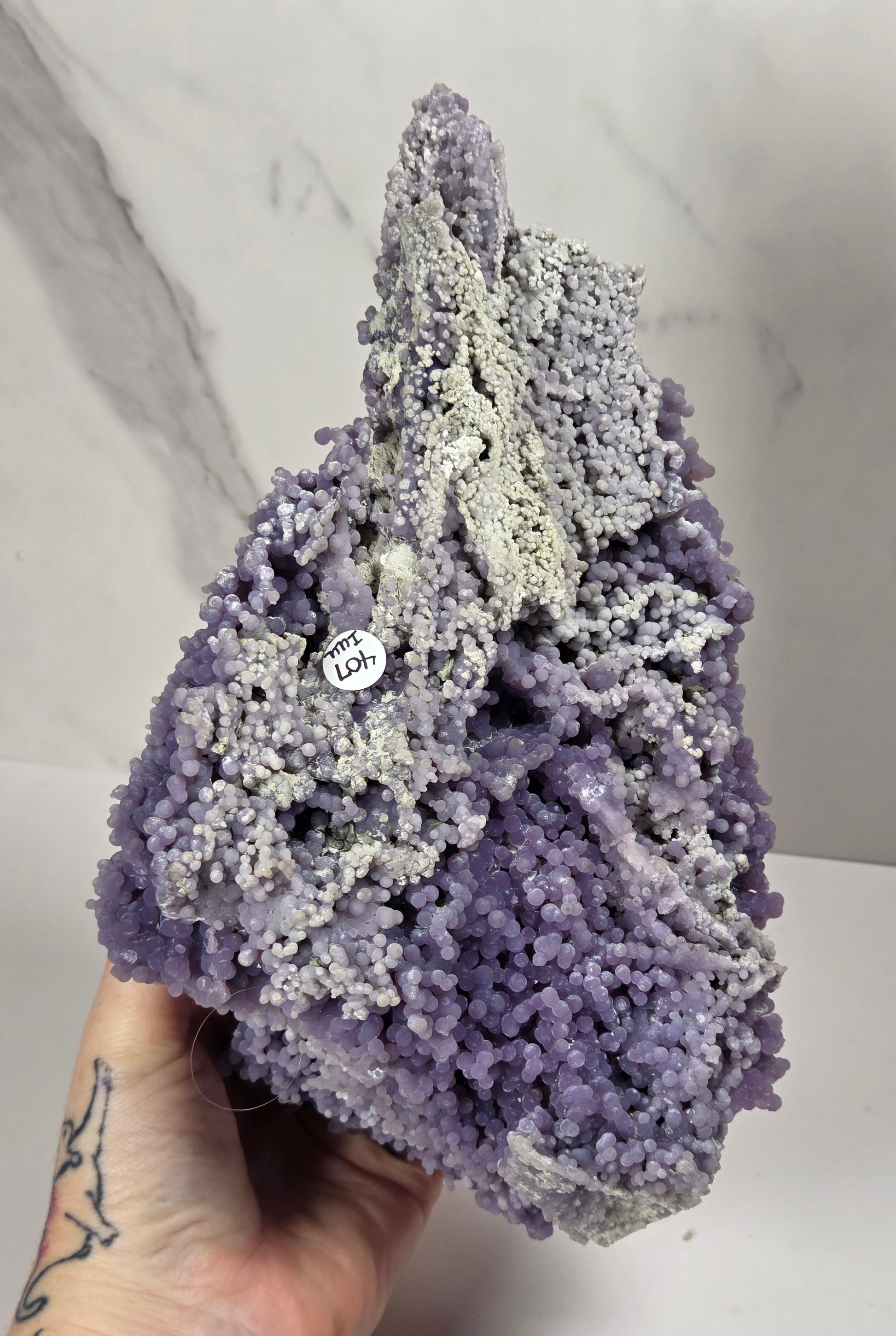 A hand holds a large purple and lavender crystalline mineral specimen with a white tag attached, set against a light gray marble-patterned background.