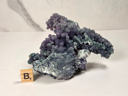 A purple and blue-green mineral specimen with a rough crystalline texture, displayed on a white surface next to a small wooden Scrabble tile showing the letter B.