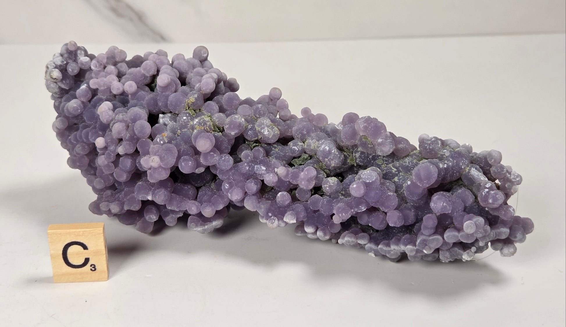 A purple amethyst cluster mineral specimen with rounded crystal balls, displayed on a white surface next to a wooden Scrabble tile showing the letter C.