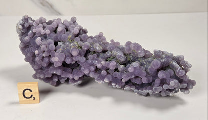 A purple amethyst cluster mineral specimen with rounded crystal balls, displayed on a white surface next to a wooden Scrabble tile showing the letter C.