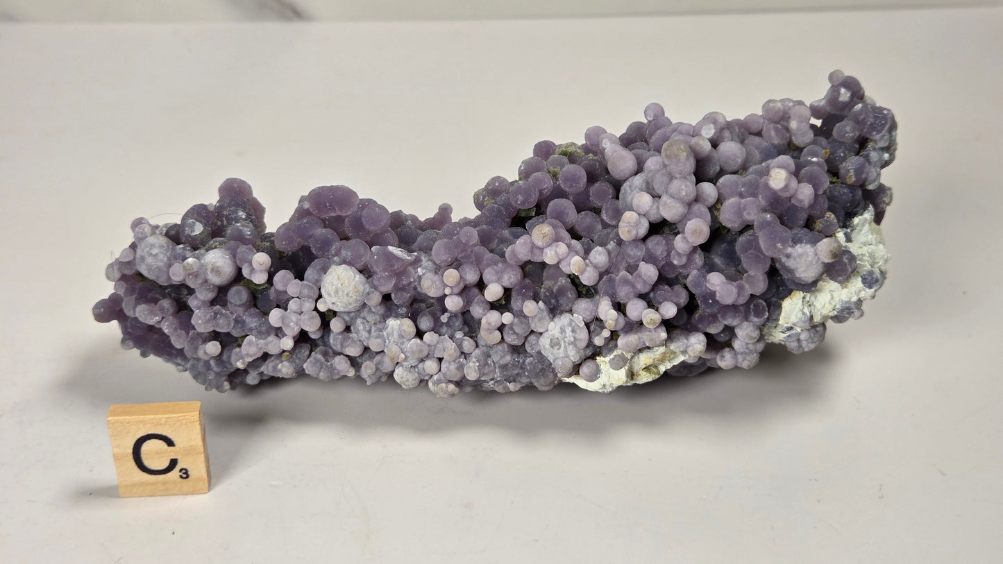 A purple and lavender clustered mineral specimen with a small wooden letter block showing 'Ca' on a white surface.