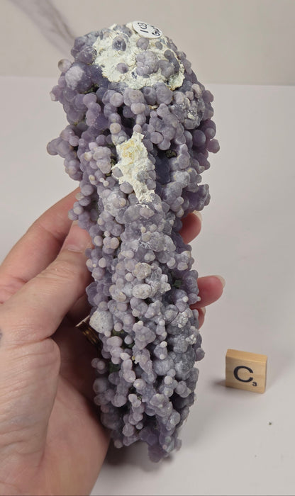A hand holding a textured lavender-purple and white mineral specimen, with a small wooden letter tile on a white tabletop.