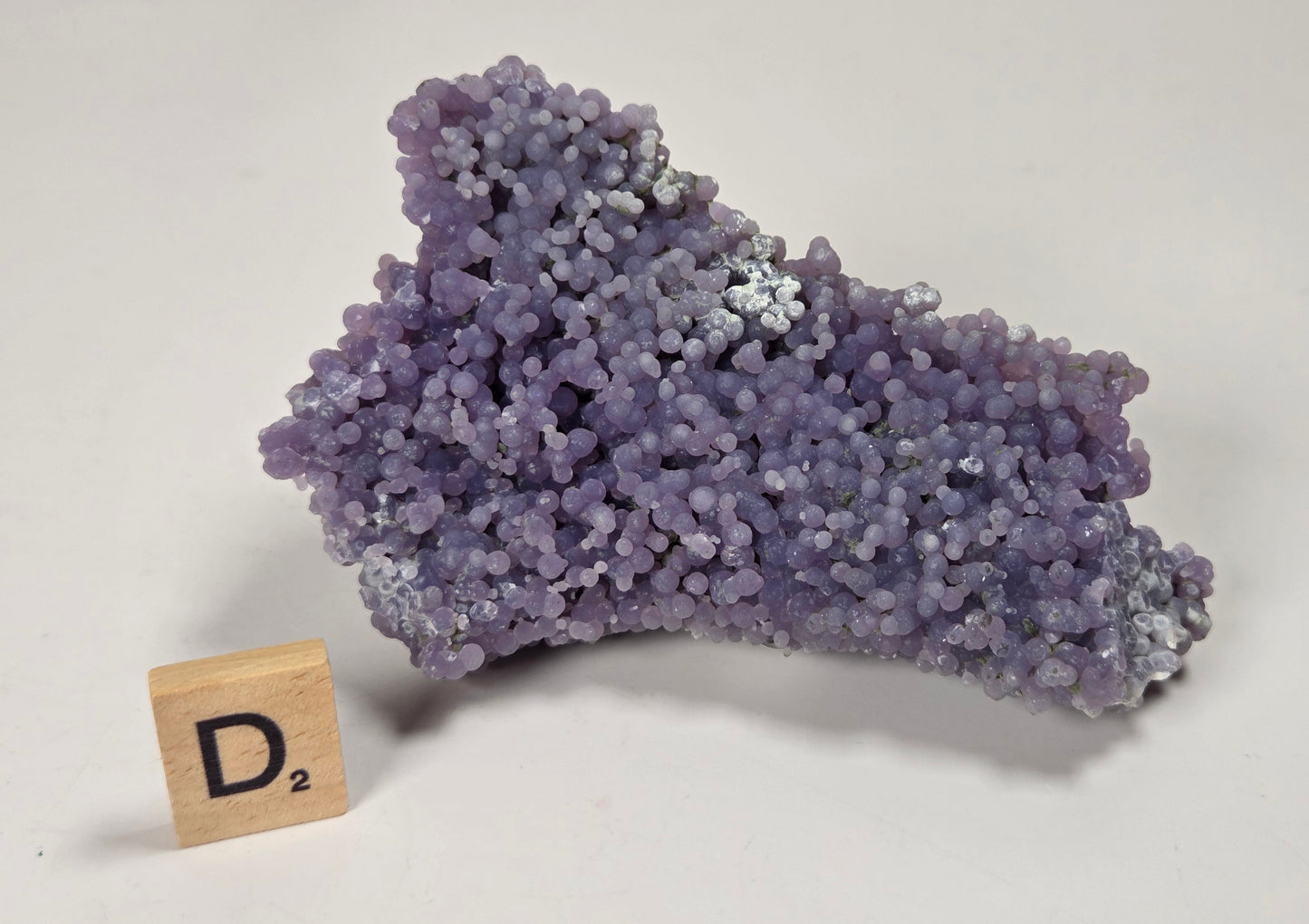 A purple crystalline mineral specimen with a rough, clustered texture, displayed on a white surface next to a small wooden letter tile showing the letter 'D'.