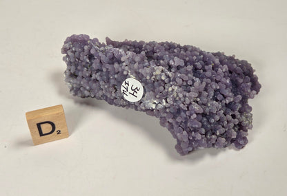 A purple crystalline mineral specimen with a rough, clustered texture, shown on a light beige background next to a small wooden block labeled 'D2'.