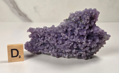 A purple amethyst crystal cluster with a small wooden Scrabble tile spelling D2 placed beside it on a light surface.