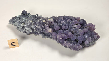 A purple and lavender crystal cluster specimen on a light gray background, with a small wooden block labeled 'E.' placed beside it for scale.