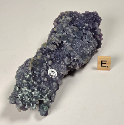 A cluster of purple and blue crystalline mineral specimen with a small white sticker reading 'JS exa' and a small wooden letter tile showing 'E.' on a white surface.