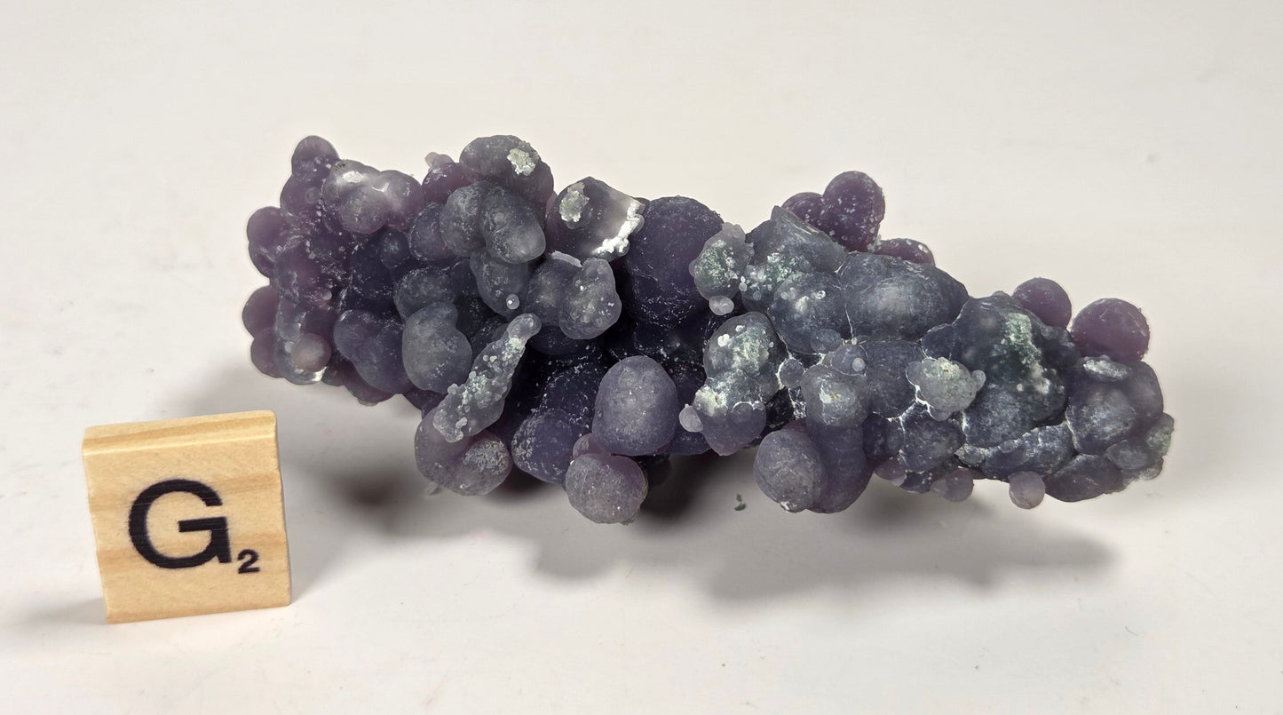 A cluster of purple grape-like beads or bumpy round objects arranged horizontally, photographed on a plain light background. A small wooden letter tile with the letter G is placed beside it for scale.