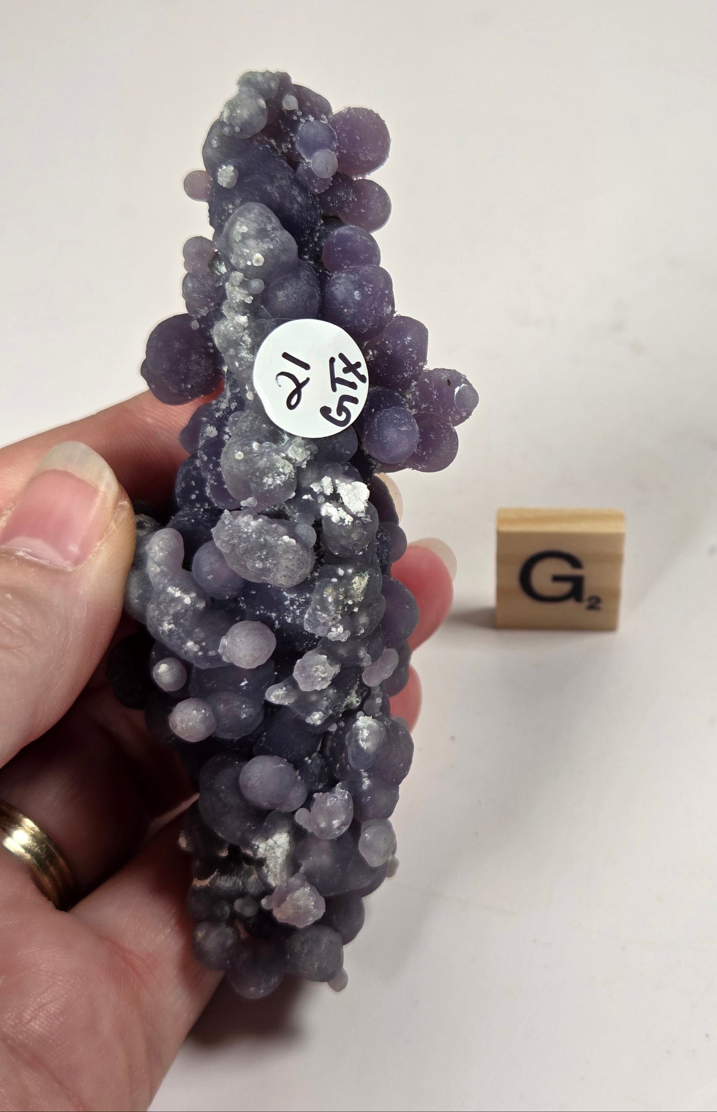 A hand holding a cluster of purple and gray grape-like gemstone beads, with a small wooden Scrabble tile in the background on a light surface.