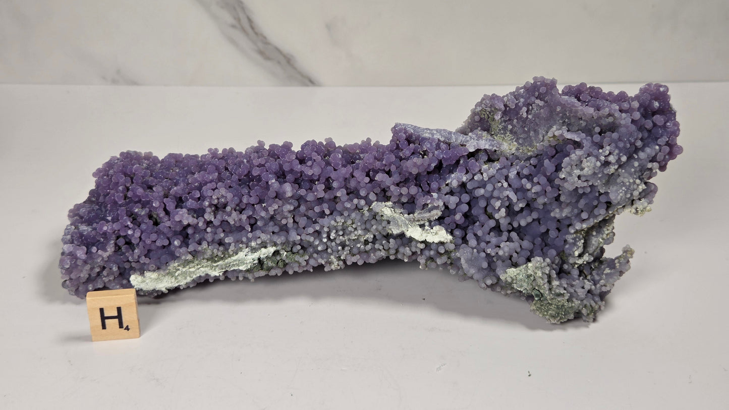A purple crystal cluster specimen, likely amethyst, displayed on a white surface with a small wooden letter block showing 'H.' in front, against a white and gray marble-patterned background.