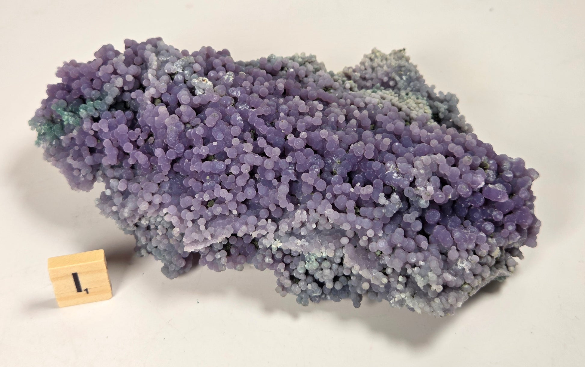 A purple and blue-gray crystalline mineral specimen with a textured, bubble-like surface, displayed on a plain off-white background with a small wooden block showing the number 1.