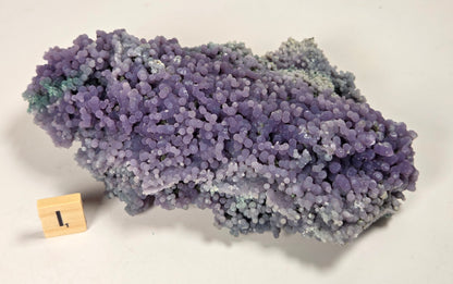 A purple and blue-gray crystalline mineral specimen with a textured, bubble-like surface, displayed on a plain off-white background with a small wooden block showing the number 1.