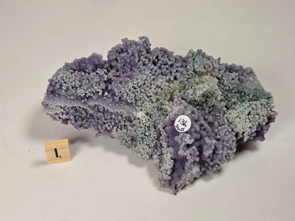 A textured mineral specimen with a purple, blue, and gray crystalline structure, displayed on a light gray background with a small wooden block marker and a white circular sticker.