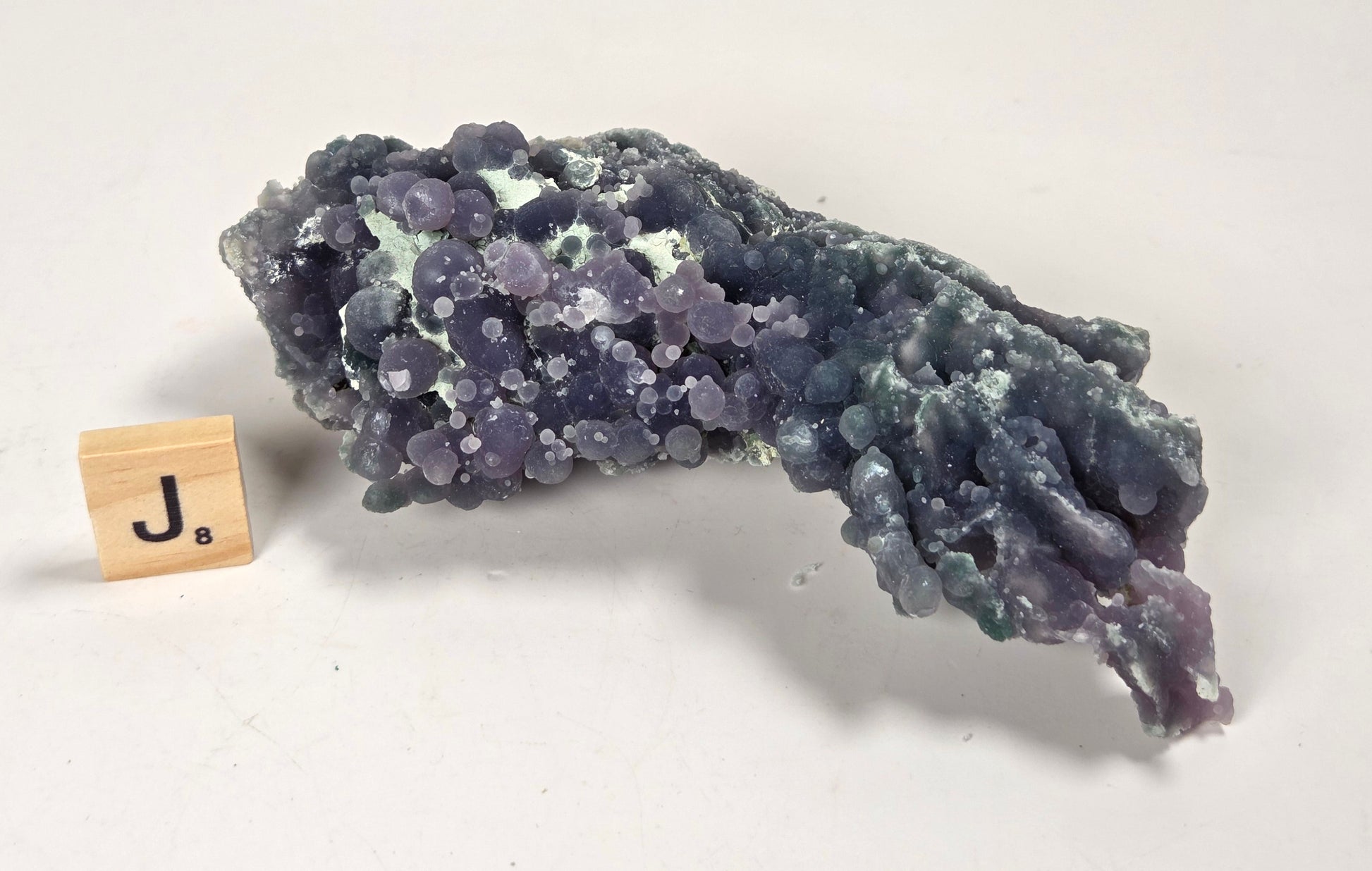 A purple and blue-green mineral specimen with a bubbly, clustered texture, displayed on a white surface next to a small wooden block with the letter J.