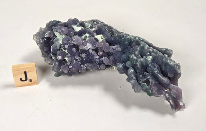 A purple and blue-green mineral specimen with a bubbly, clustered texture, displayed on a white surface next to a small wooden block with the letter J.
