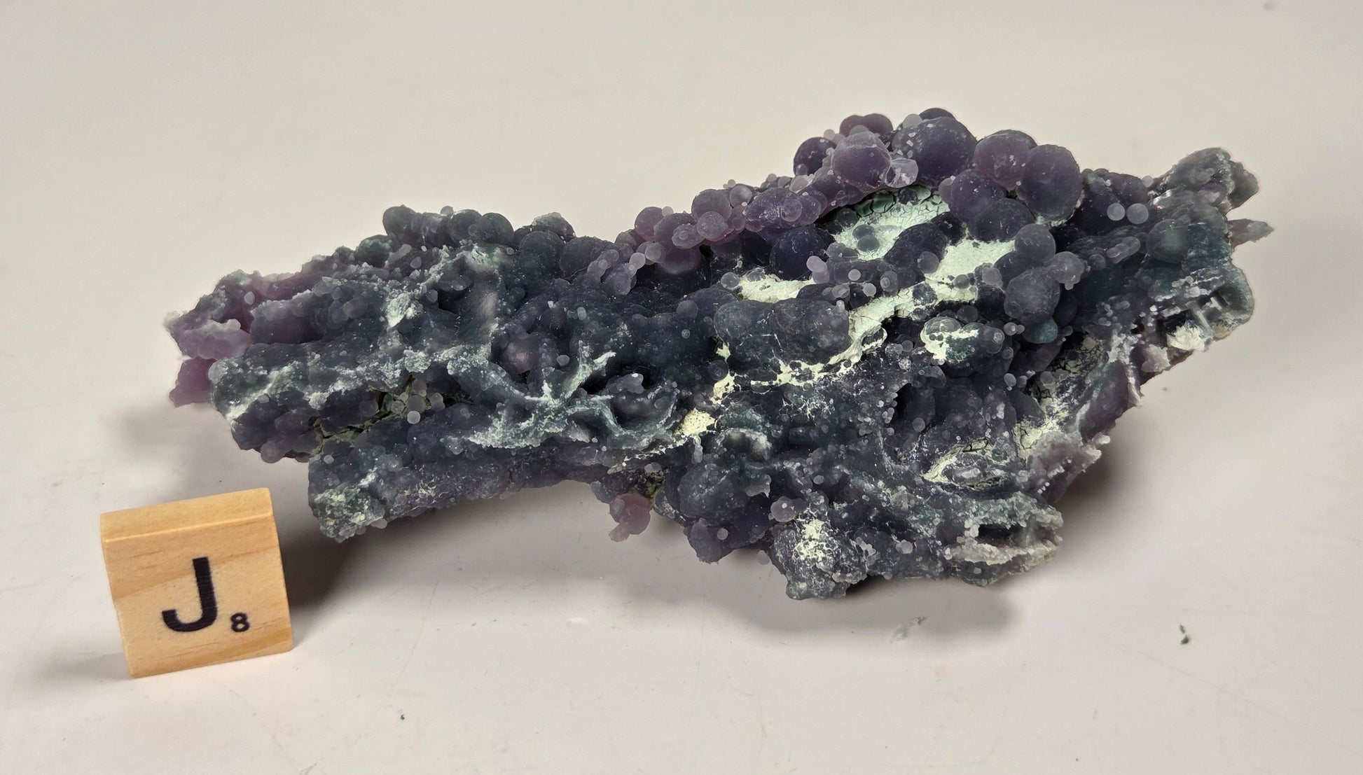 A natural purple and blue-gray mineral specimen with a rough, clustered texture, displayed on a light beige background next to a small wooden Jenga block.