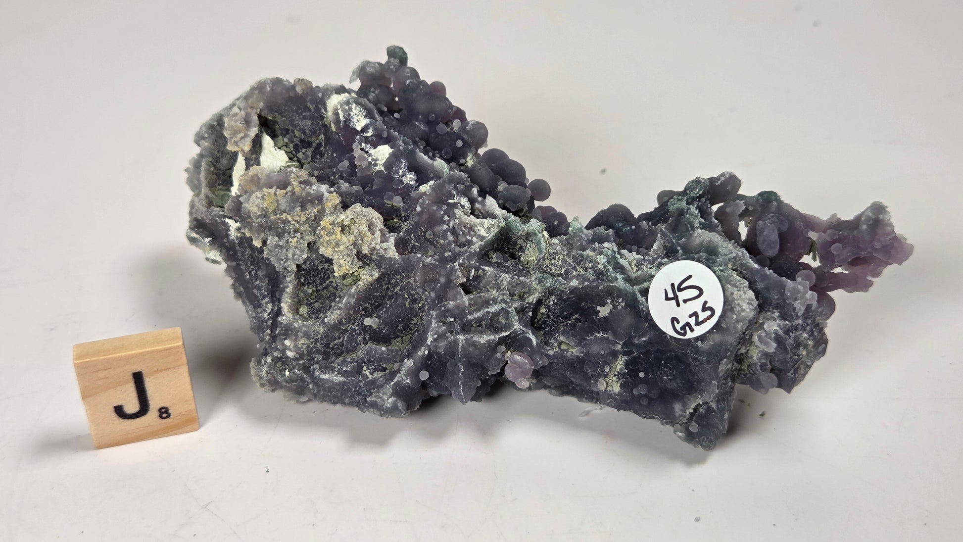 A purple and gray mineral specimen with a rough, crystalline texture, displayed on a light gray background next to a small wooden tile with a 'J' and a white inventory sticker.